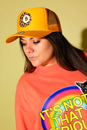 A woman wearing a yellow trucker hat with a leopard print and number 8 patch, paired with a bright orange graphic sweatshirt, set against a solid pale yellow background.