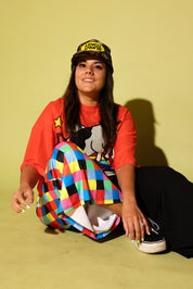 Model sitting wearing an orange mesh top with a cow and rainbow graphic and colorful pants on a yellow background
