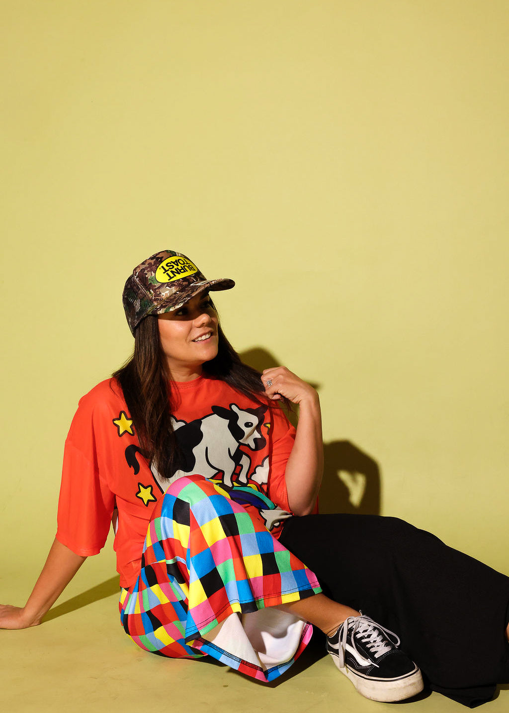 A woman sitting on a yellow-green studio floor wearing a bright orange graphic t-shirt, a colorful plaid skirt, black pants, and a camouflage baseball cap with a yellow logo patch.