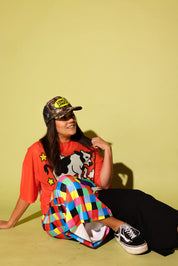 A woman sitting on a yellow-green studio floor wearing a bright orange graphic t-shirt, a colorful plaid skirt, black pants, and a camouflage baseball cap with a yellow logo patch.