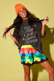Model wearing a grey graphic tee and colorful checkered satin shorts and an orange beanie on a pale yellow background