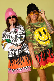 Two women posing in streetwear outfits against a solid pale yellow-green background. The woman on the left wears a pink beanie, black sunglasses, a black-and-white cow-print jacket, and an orange skirt with a black drips pattern and pink lace trim.
