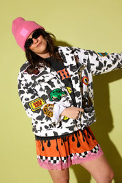 Model wearing a cow pattern jacket and bright shorts and a pink beanie on a pale yellow background