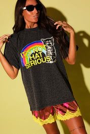 A woman modeling an oversized black graphic t-shirt with a rainbow and a cat illustration that reads 'IT'S NOT THAT SERIOUS'. She is wearing pink and yellow satin shorts against a yellow background.