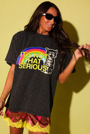 A woman wearing a black graphic t-shirt with a rainbow and a cat illustration that reads 'IT'S NOT THAT SERIOUS' in yellow text, paired with pink and yellow shorts, set against a solid yellow background.