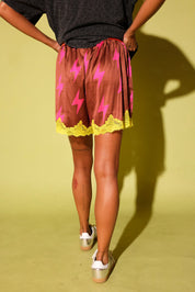 Back side of a model wearing maroon shorts with pink lightening bolts and yellow lace trim and a leopard shirt against a pale yellow background