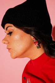 A model wearing a red crewneck and black beanie with earrings in the shape of a nutcracker