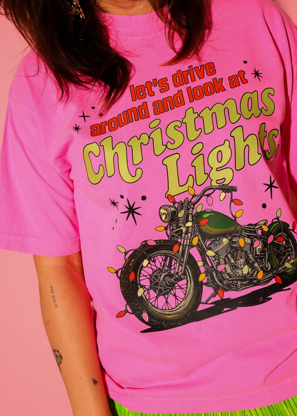 A model wearing the “let’s drive around and look at Christmas Lights.” tee in hot pink, styled with lime green plissé wide-leg pants against a pink background — graphic detail shown up close from the waist up.