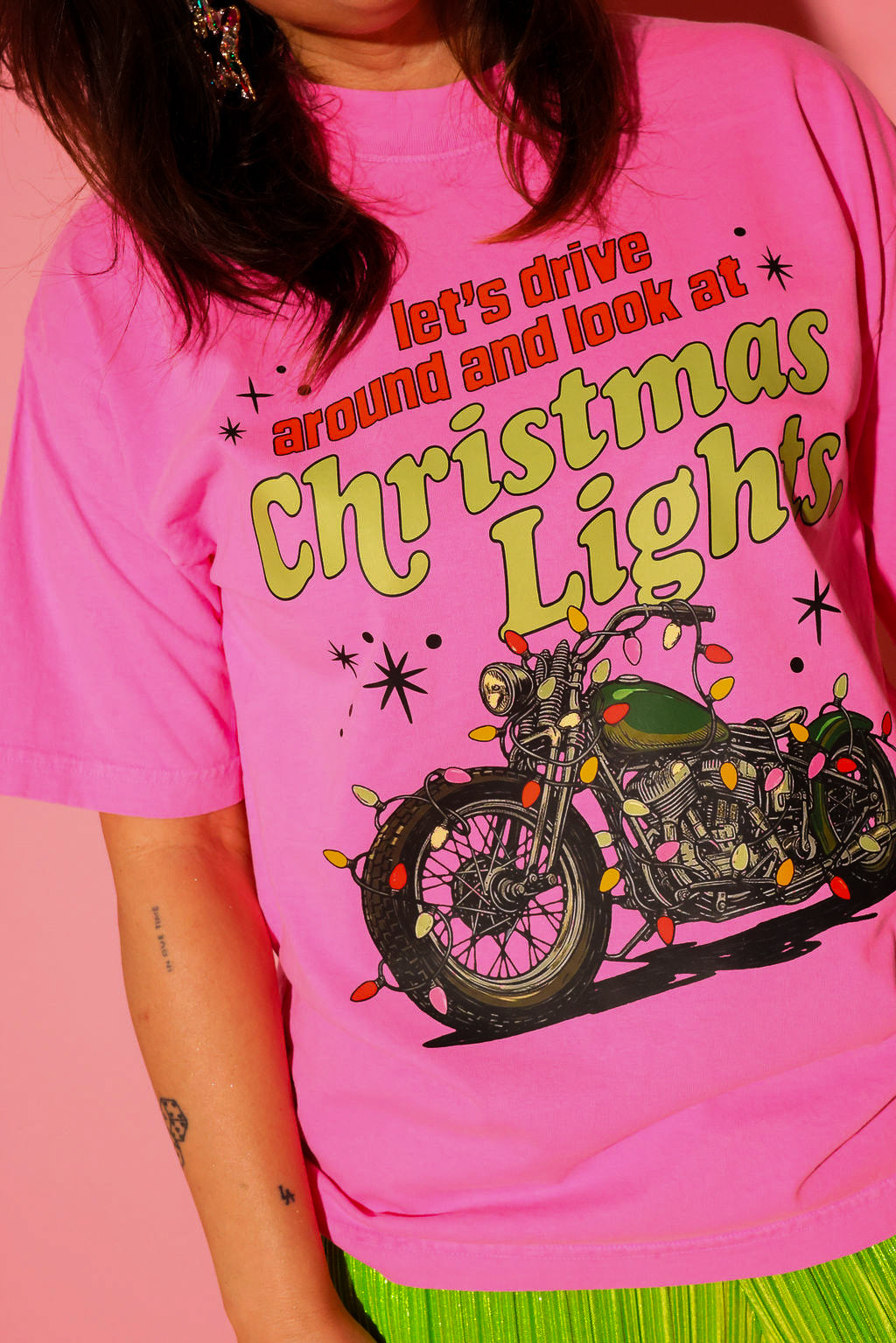 A model wearing the “let’s drive around and look at Christmas Lights.” tee in hot pink, styled with lime green plissé wide-leg pants against a pink background — graphic detail shown up close from the waist up.