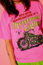 A model wearing the “let’s drive around and look at Christmas Lights.” tee in hot pink, styled with lime green plissé wide-leg pants against a pink background — graphic detail shown up close from the waist up.