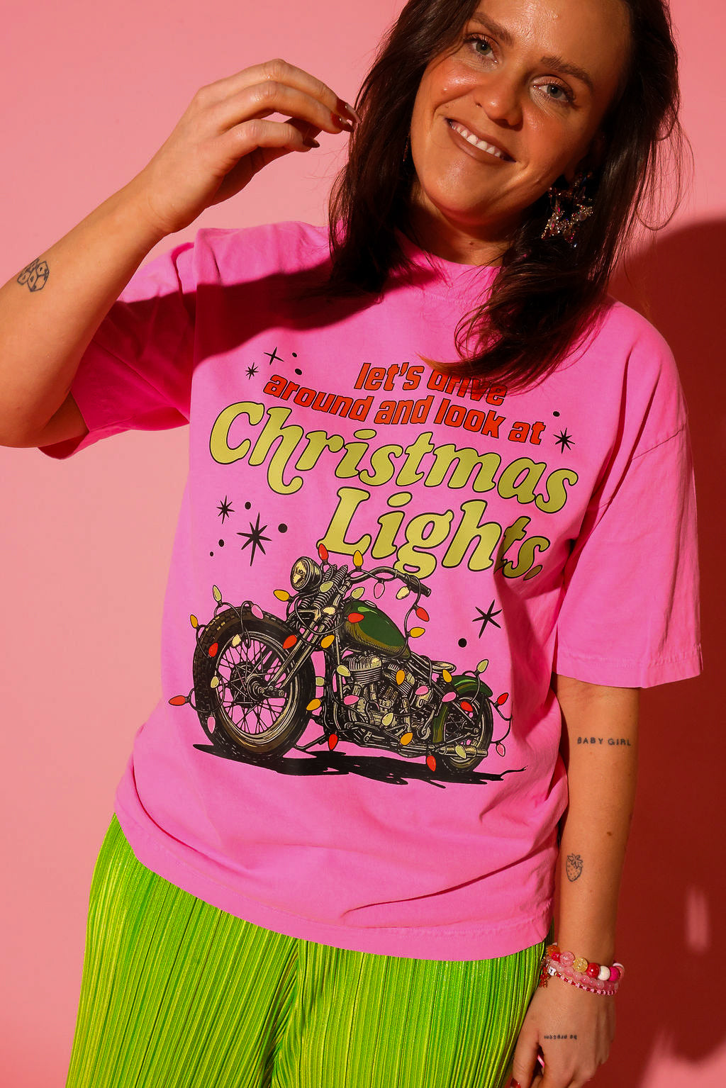 Model wearing a pink short sleeve graphic tee with text and a motorcycle covered in Christmas lights graphic, green plisse metallic pants and some jewelry, on a pink background