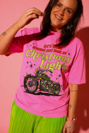 Model wearing a pink short sleeve graphic tee with text and a motorcycle covered in Christmas lights graphic, green plisse metallic pants and some jewelry, on a pink background