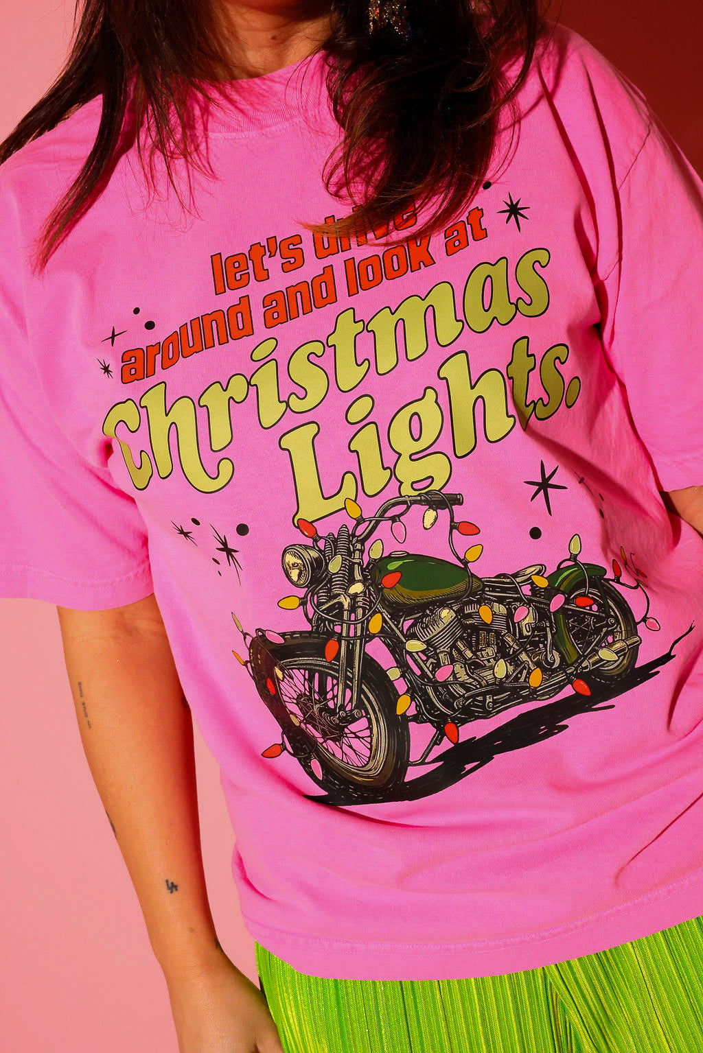 A model wearing the “let’s drive around and look at Christmas Lights.” tee in hot pink, styled with lime green plissé wide-leg pants against a pink background — graphic detail shown up close from the waist up.