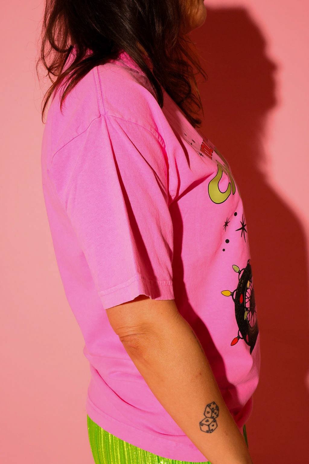 Side view of a model wearing the “let’s drive around and look at Christmas Lights.” tee in hot pink, styled with lime green plissé wide-leg pants against a pink background — graphic detail shown up close from the waist up.