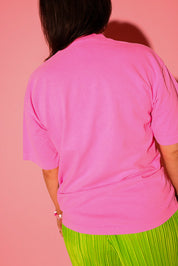 Back view of a model wearing a tee in hot pink, styled with lime green plissé wide-leg pants against a pink background
