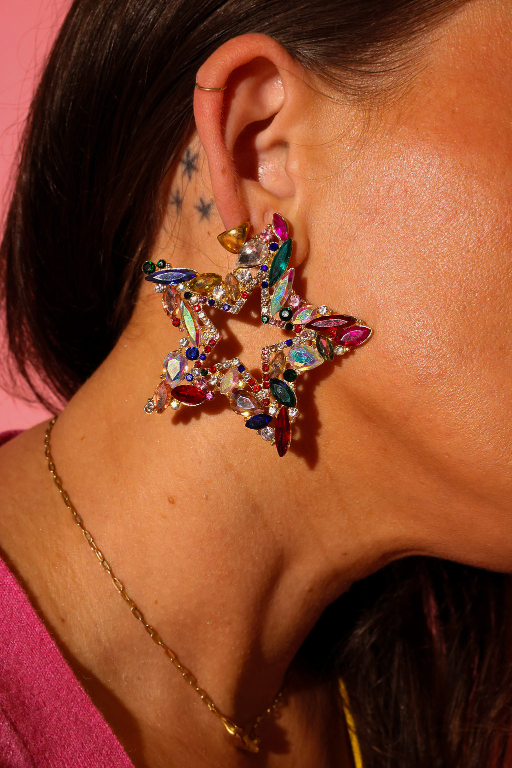 Side view of a model wearing earrings that are a large star with different color jewels, on a pink background