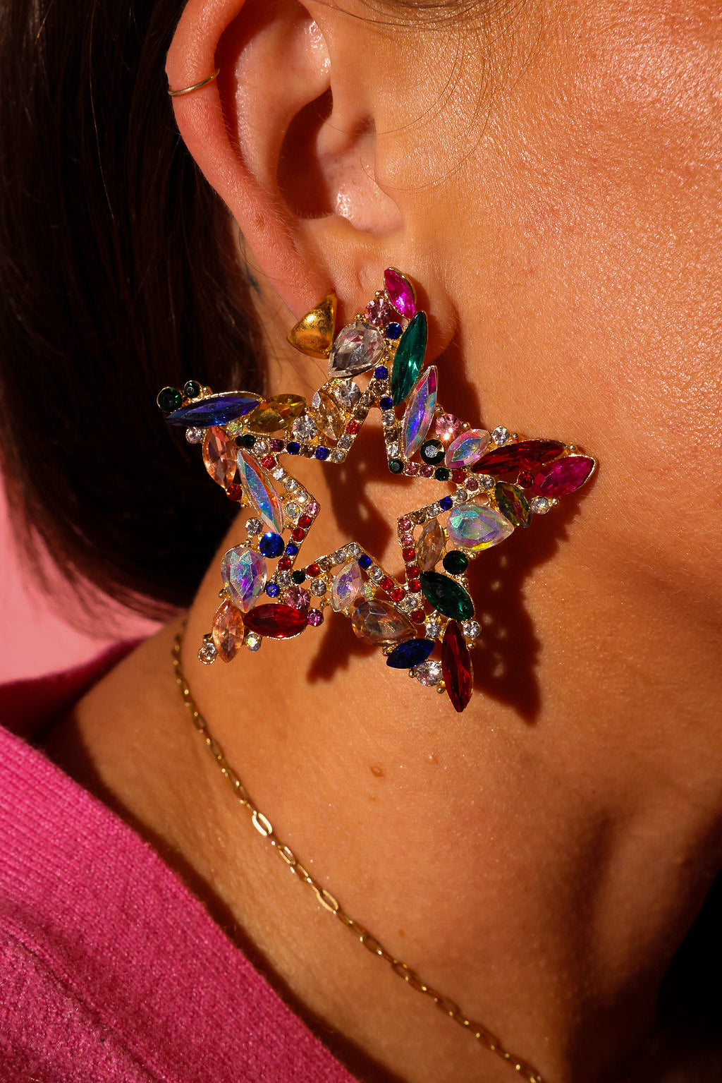 Side view of a model wearing earrings that are a large star with different color jewels, on a pink background