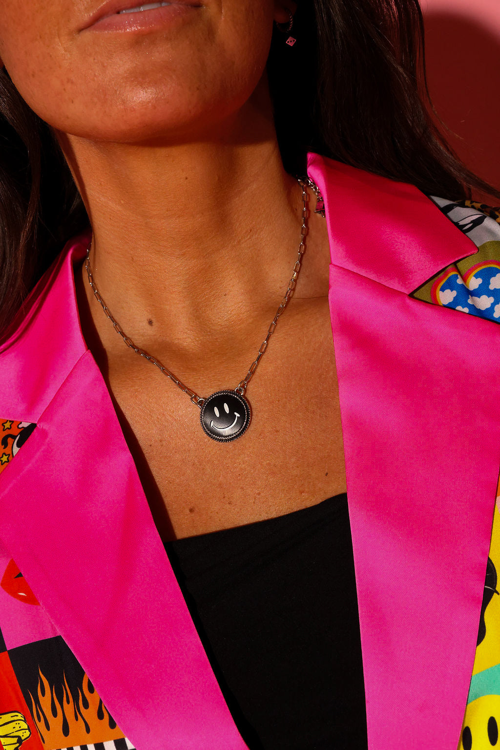 Model wearing a bright pink blazer and silver necklace with a black smiley face charm