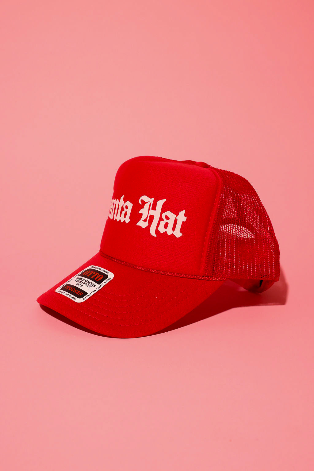 Side view of a red trucker hat with white text that reads 'Santa Hat' on a pink background
