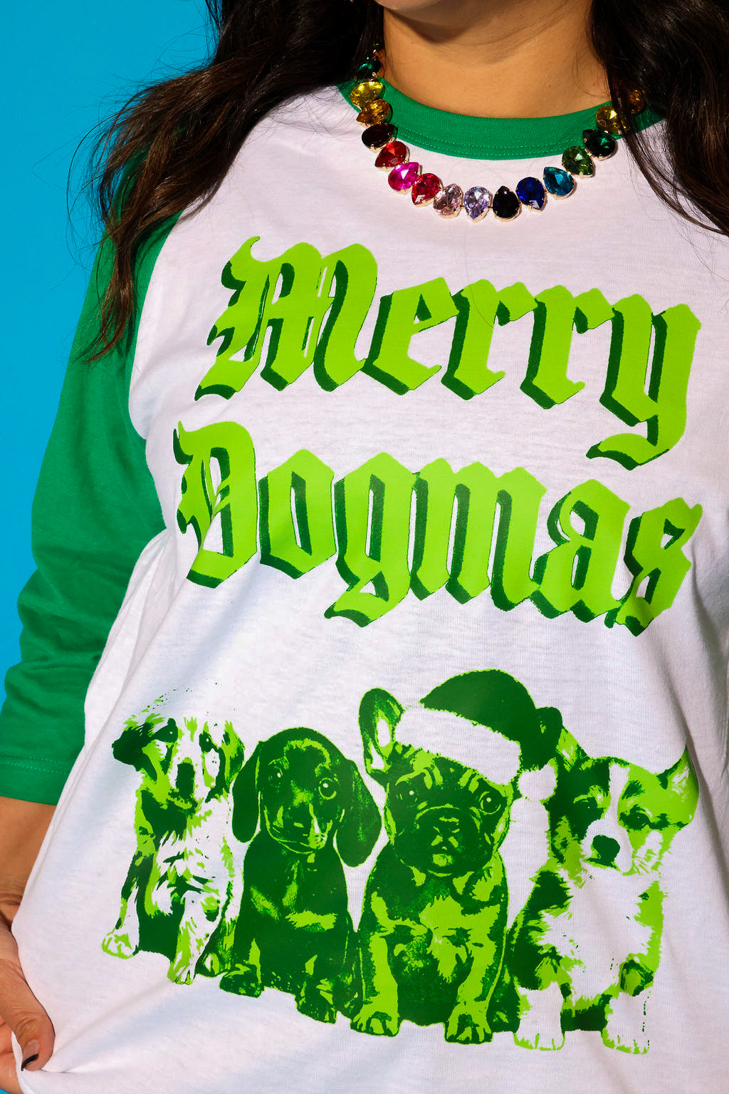 Model wearing a green baseball tee with a Christmas graphic and a rainbow jeweled necklace on a blue background