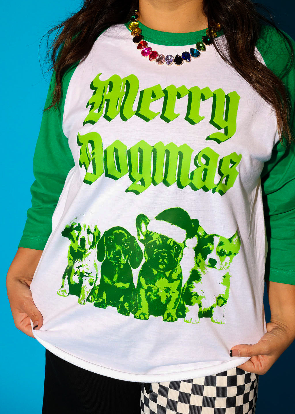 4 sleeves and a green collar, featuring “Merry Dogmas” in bold old English lettering above four illustrated puppies in a tonal green duotone print, one wearing a Santa hat. Styled with a multicolor crystal necklace and black and white checkerboard pants.