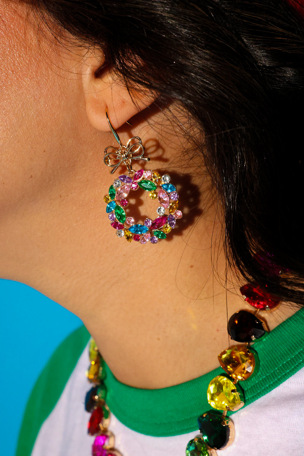 Close up side view of a model wearing jeweled wreath earrings and necklace and a green baseball tee on a blue background