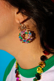 Close up side view of a model wearing jeweled wreath earrings and necklace and a green baseball tee on a blue background