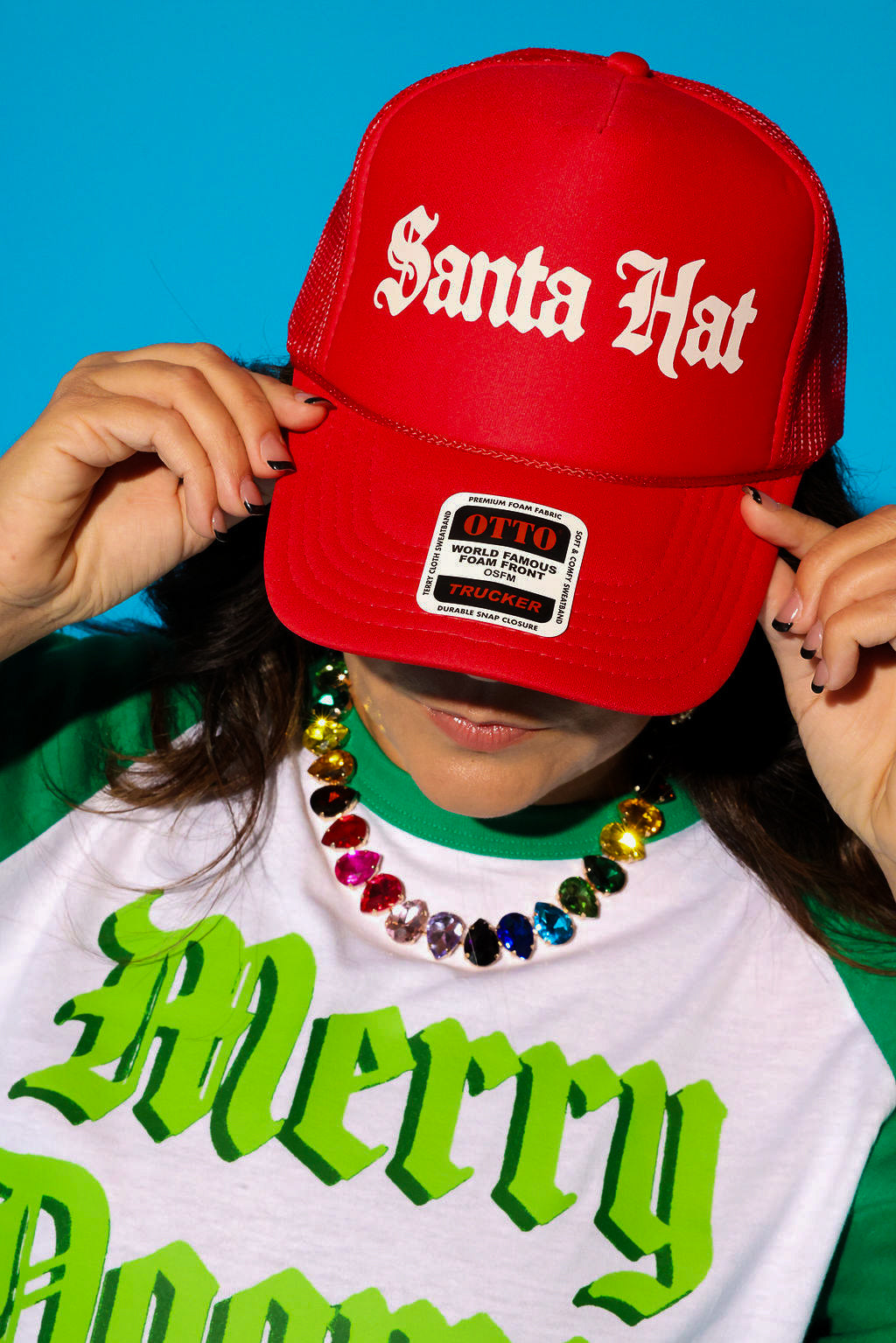 A close-up of a model tilting a red trucker hat that reads “Santa Hat” in cream gothic lettering down over her face, styled with a white and green “Merry Dogmas” graphic tee layered over a green crewneck and a rainbow crystal statement necklace. Background is light blue.