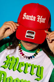 A close-up of a model tilting a red trucker hat that reads “Santa Hat” in cream gothic lettering down over her face, styled with a white and green “Merry Dogmas” graphic tee layered over a green crewneck and a rainbow crystal statement necklace. Background is light blue.