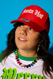 A close-up of a model tilting a red trucker hat that reads “Santa Hat” in cream gothic lettering down over her face, styled with a white and green “Merry Dogmas” graphic tee layered over a green crewneck and a rainbow crystal statement necklace. Background is light blue.