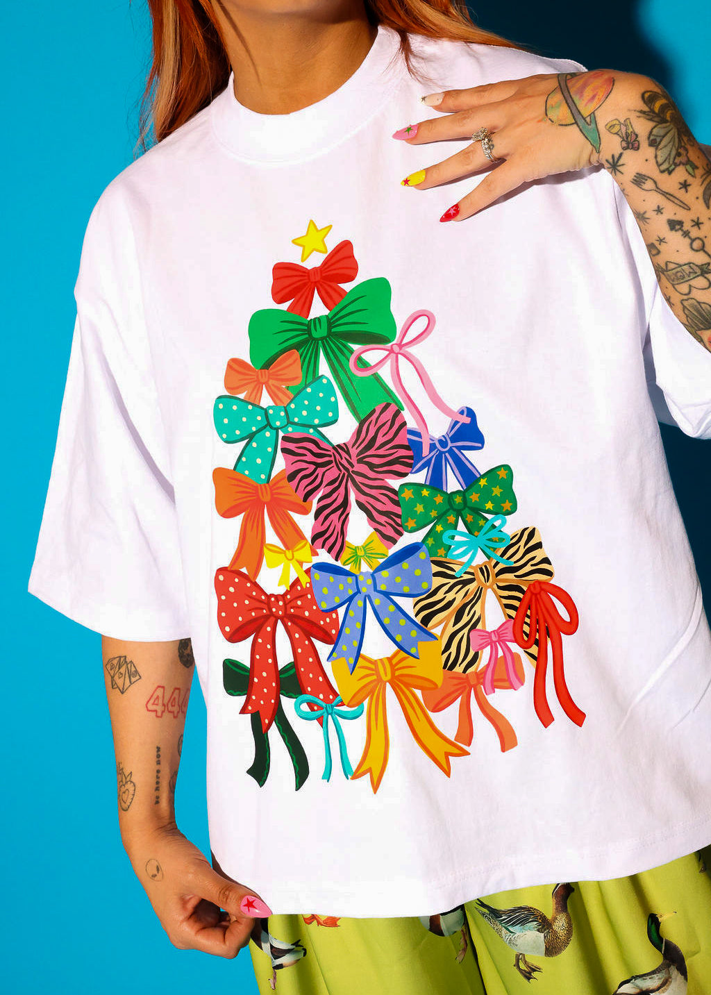 Close-up detail shot of the bow Christmas tree graphic on the white oversized tee, worn by a model with red hair and heavily tattooed arms, styled with yellow-green duck print pants against a blue background.