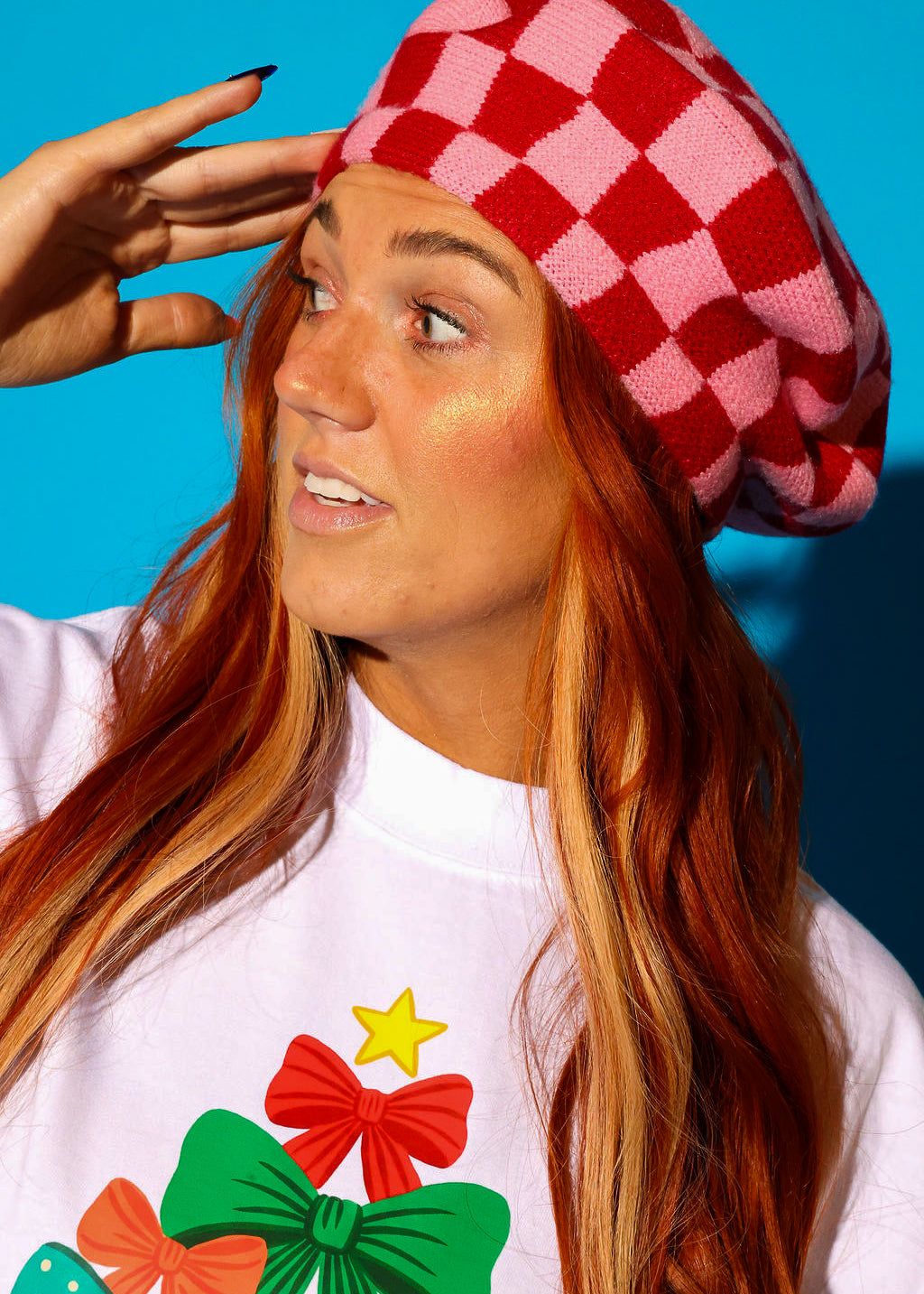 Close up of a model wearing a pink and red checkered beret and white graphic tee with holiday bows on a blue background