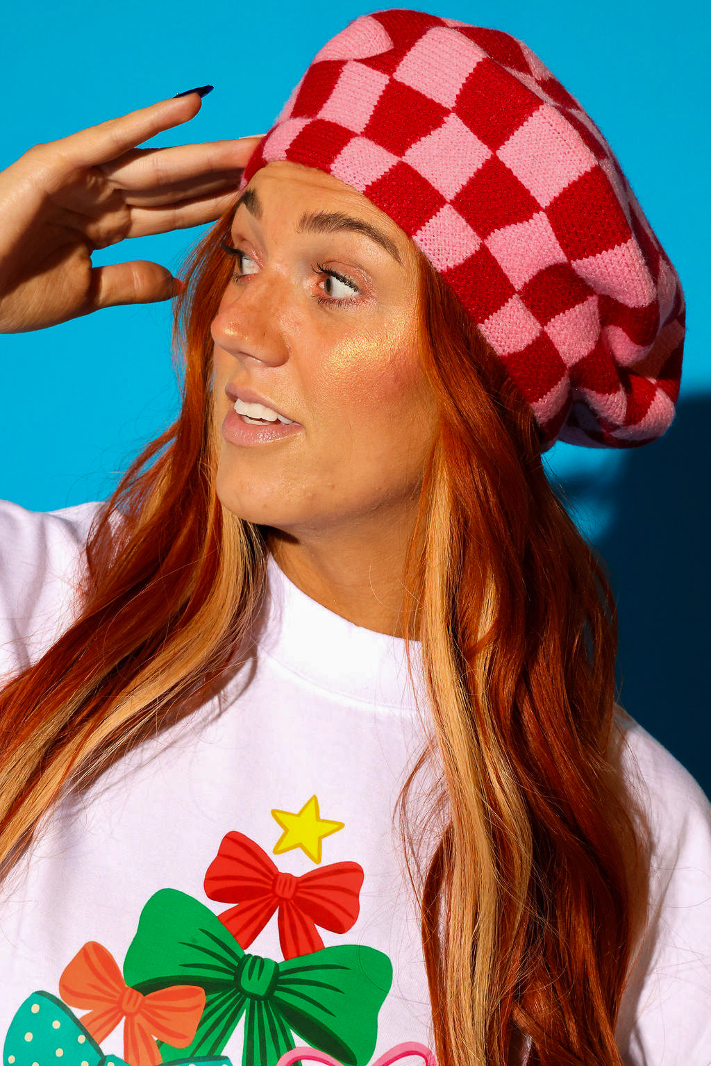 Close up of a model wearing a pink and red checkered beret and white graphic tee with holiday bows on a blue background