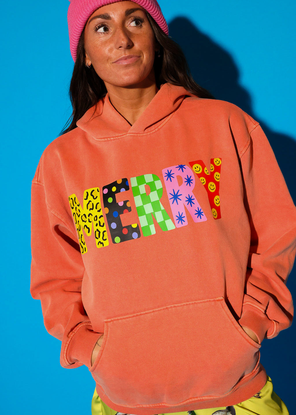 A model wearing the washed coral “MERRY” hoodie styled with yellow-green duck print lace-trim shorts against a blue background — close-up detail shot showing the multicolor patterned block letter graphic clearly across the chest.