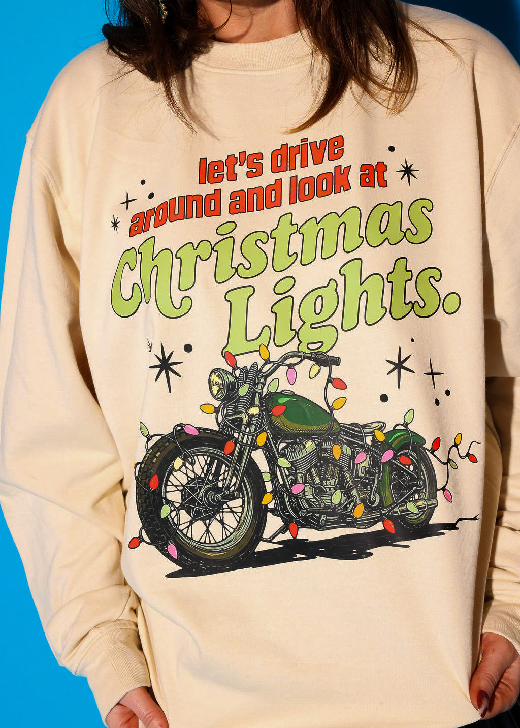 Close-up detail shot of the “let’s drive around and look at Christmas Lights.” graphic on the cream sweatshirt, showing the full retro motorcycle and text design in vivid detail against a blue background.