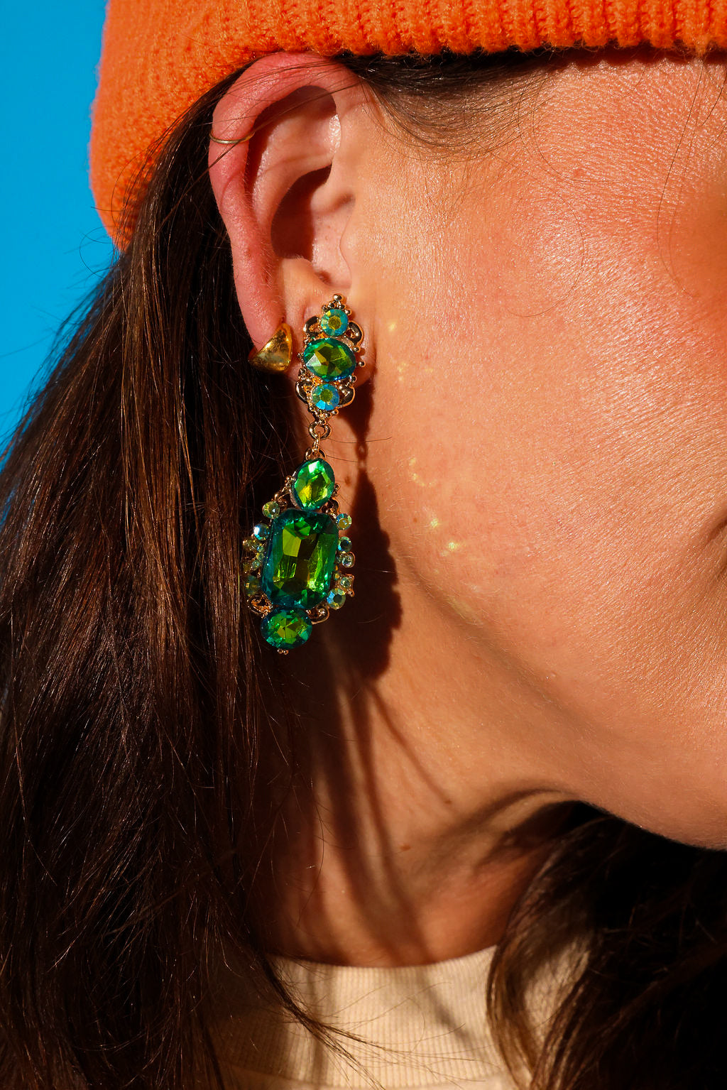 Close up side view of a model wearing green dangly earrings and an orange beanie on a blue background