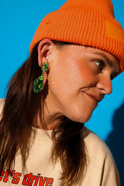 Close up side view of a model wearing green dangly earrings, cream shirt, and an orange beanie on a blue background