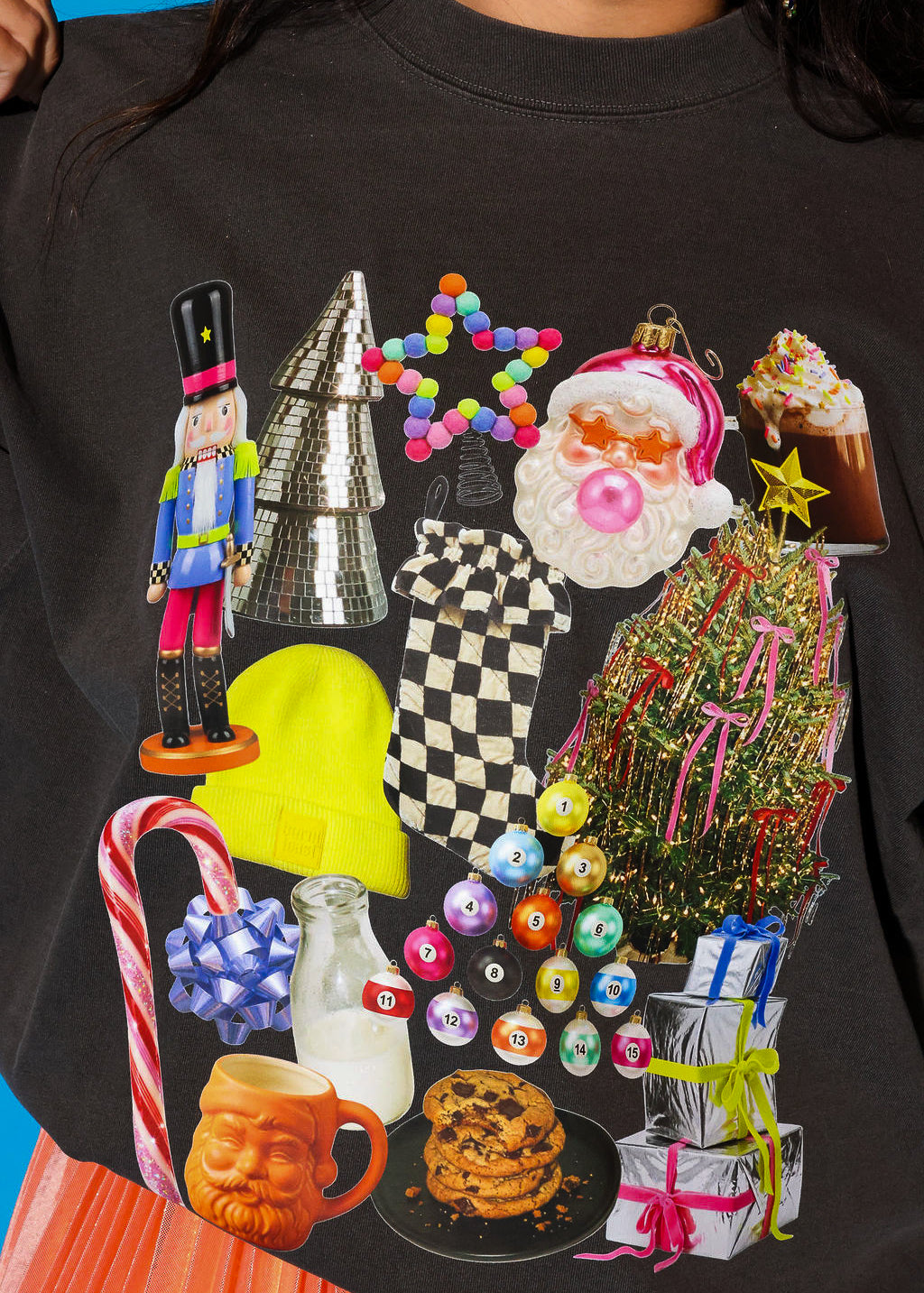 Close-up detail shot of the Holiday Collage graphic on a charcoal washed crewneck sweatshirt, showing the full maximalist holiday collage print up close — worn by a model with an orange plissé skirt visible at the hem and a blue background peeking in at the sides.