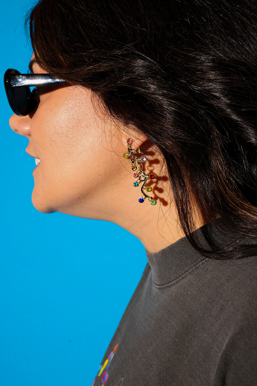 Model wearing a grey tee sunglasses and dangly earrings with multi-colored jewels on a blue background