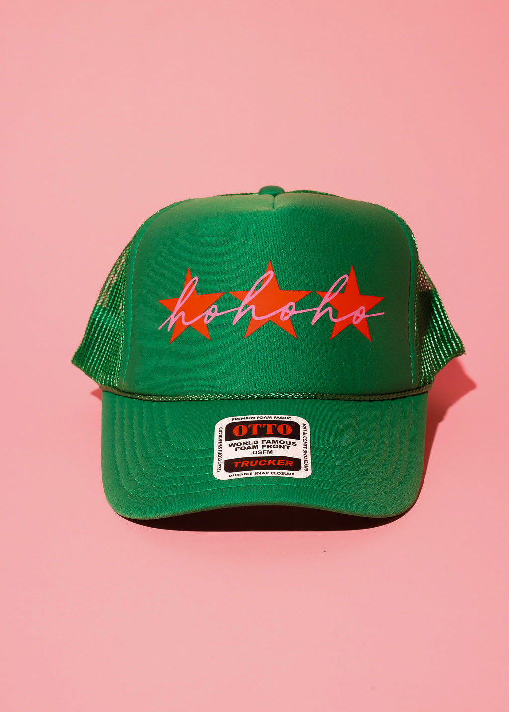 A green trucker hat with three red stars and cursive text in pink that reads 'ho ho ho' on a pink background