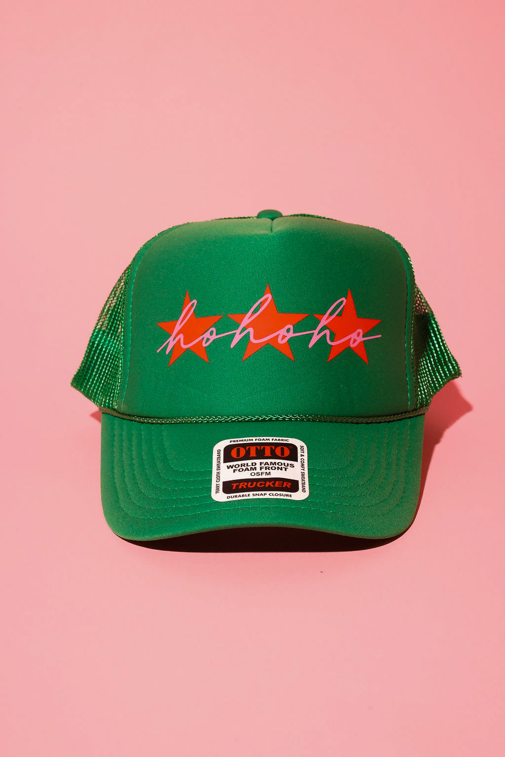 A green trucker hat with three red stars and cursive text in pink that reads 'ho ho ho' on a pink background