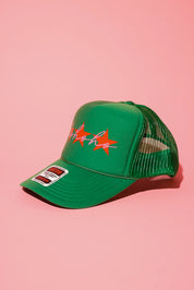 Side view of a green trucker hat with three red stars and cursive text in pink that reads 'ho ho ho' on a pink background