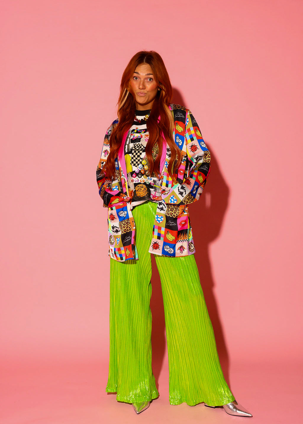 A model stands against a pink background wearing the One Year of Burnt Toast Blazer in bold multicolor patchwork print with pink satin lapels, styled over a black and white striped top with lime green metallic wide-leg plissé pants and silver pointed-toe heels.