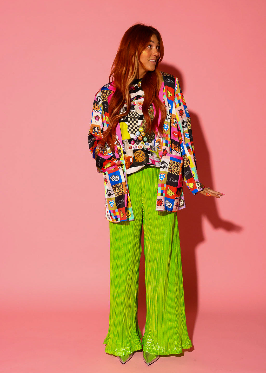 A model wearing an oversized blazer covered in a colorful Burnt Toast patchwork print featuring smileys, leopard, flames, bananas, dice, ladybugs, orcas, 8-balls, black panthers, and “I ❤️ Burnt Toast” graphics, with pink satin lapels and cuffs. Styled over a holiday collage graphic tee and lime green pleated pants, on a pink background