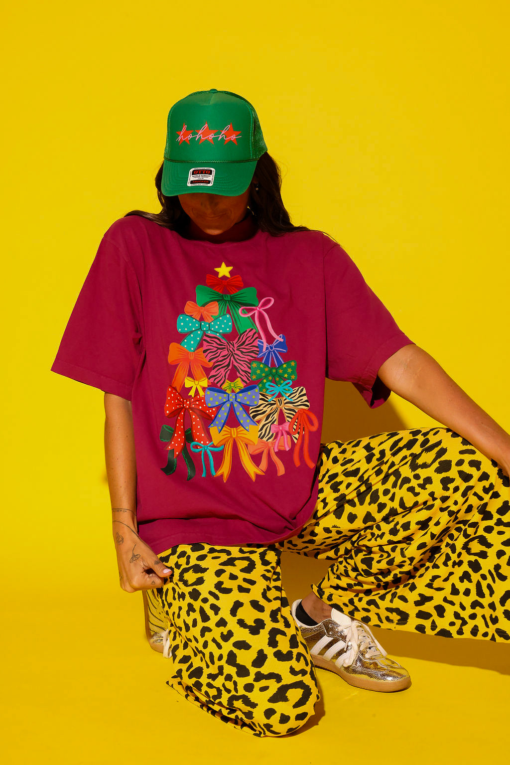 A model crouches against a yellow background wearing the Tied With a Bow graphic tee in burgundy featuring a colorful bow Christmas tree, yellow leopard print wide-leg pants, a green “Ho Ho Ho” trucker hat, and gold Adidas sneakers.