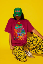 A model crouches against a yellow background wearing the Tied With a Bow graphic tee in burgundy featuring a colorful bow Christmas tree, yellow leopard print wide-leg pants, a green “Ho Ho Ho” trucker hat, and gold Adidas sneakers.