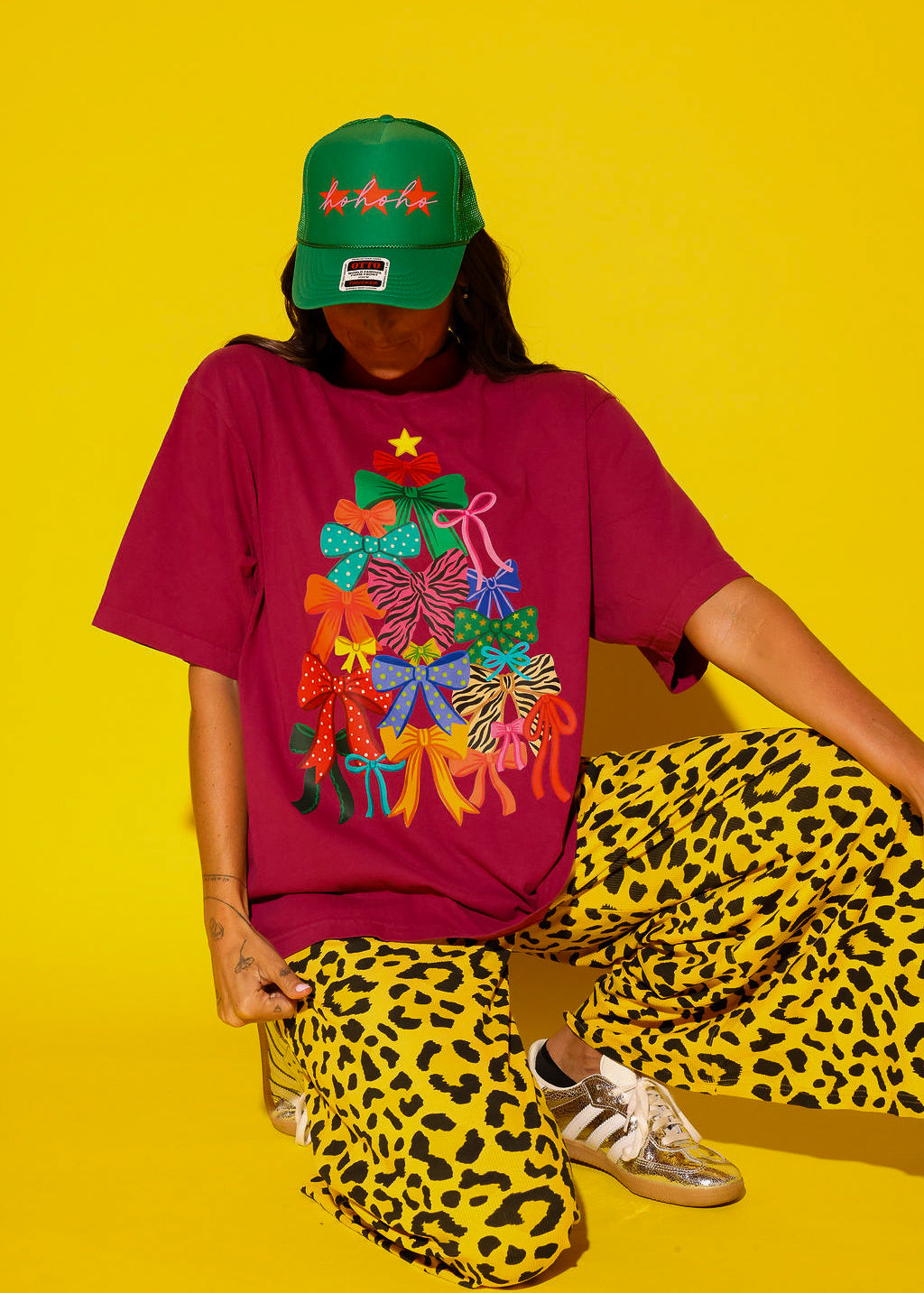 A model crouches against a yellow background wearing a burgundy oversized tee with a colorful bow Christmas tree graphic, paired with yellow leopard print wide-leg pants, a green “ho ho ho” trucker hat, and gold sneakers.