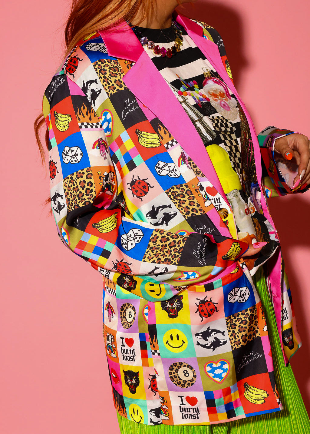 A model wearing an oversized blazer covered in a colorful Burnt Toast patchwork print featuring smileys, leopard, flames, bananas, dice, ladybugs, orcas, 8-balls, black panthers, and “I ❤️ Burnt Toast” graphics, with pink satin lapels and cuffs. Styled over a holiday collage graphic tee and lime green pleated pants.