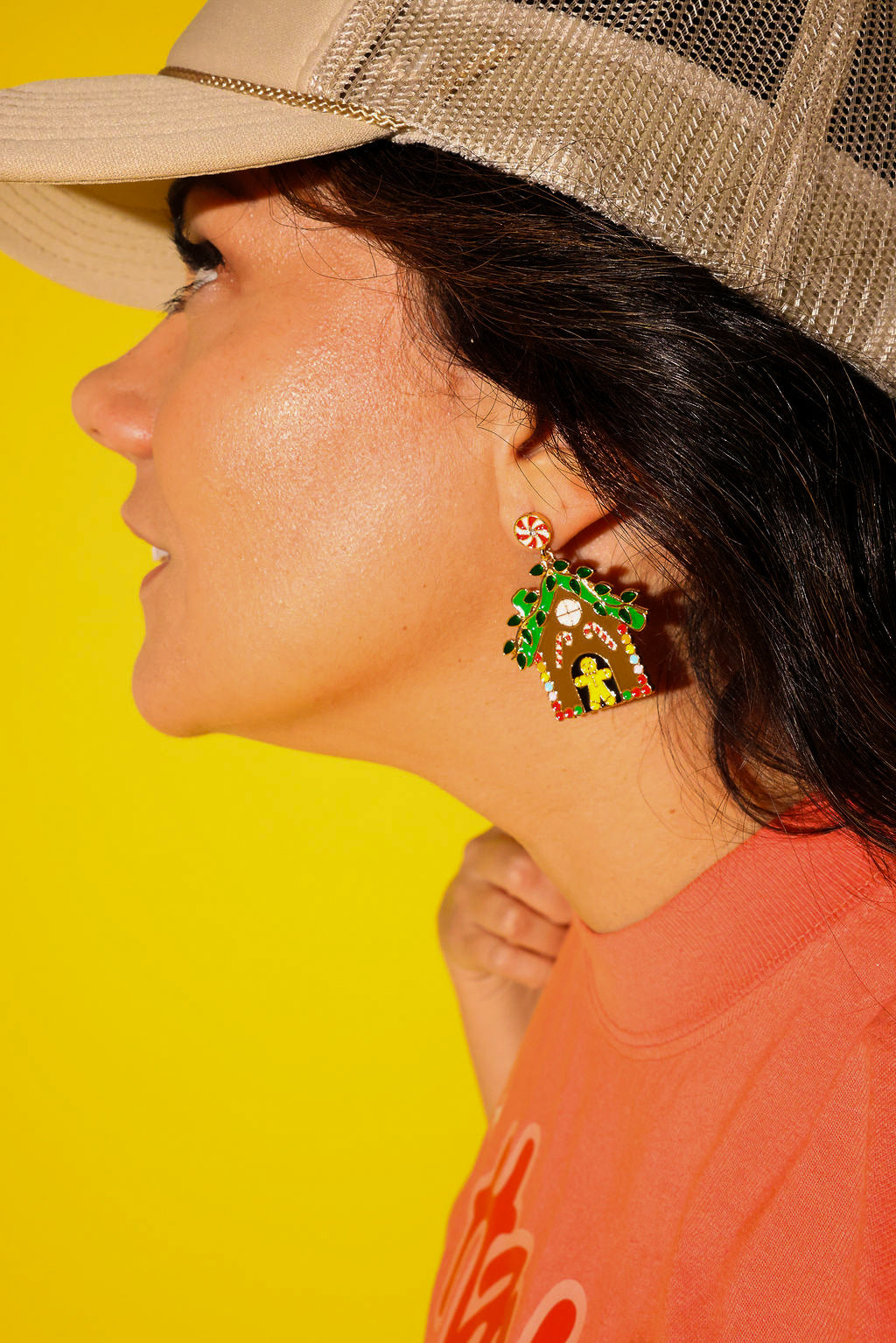 Close up side view of a model wearing an orange tee, cream trucker hat, and gingerbread earrings on a yellow background