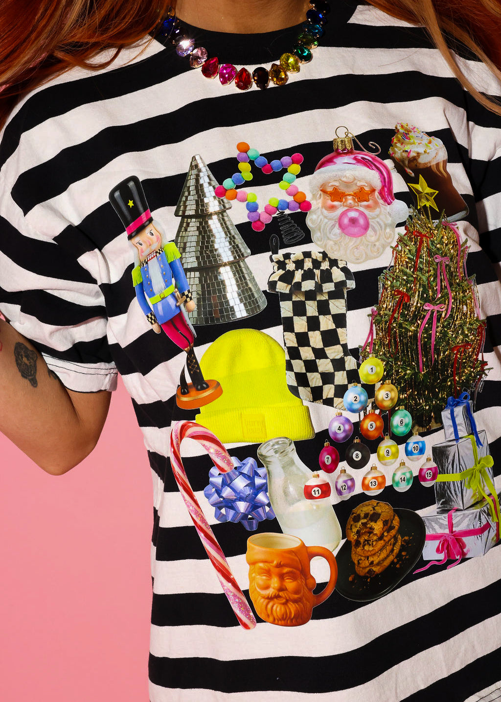 Model wearing a black and white striped tee with a Christmas graphic and a rainbow jeweled necklace on a pink background